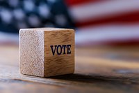 Wooden cube with VOTE text against blurred American flag background on rustic wooden surface, symbolizing democracy and electoral process.