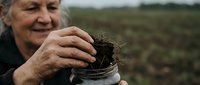 Senior Caucasian male farmer examining rich dark soil sample in glass jar outdoors, sustainable agriculture and earth quality testing concept.