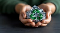 Female hands holding raw emerald crystal in silver foil against dark background, showcasing natural gemstone mineral specimen for jewelry or collection.