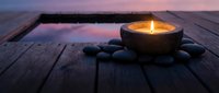 Zen spa candle with smooth stones on wooden deck at sunset creating peaceful meditation atmosphere for wellness and relaxation rituals.