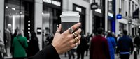 Closeup of hand holding white takeaway coffee cup on busy urban street with blurred pedestrians and storefronts in background.