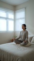 Asian woman practicing morning meditation in peaceful bedroom with natural light streaming through corner windows for mindfulness and wellness routine.