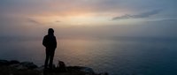 Silhouetted figure standing on rocky cliff overlooking calm ocean at sunset, contemplative mood for travel, meditation, and lifestyle concepts.