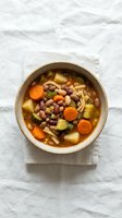 Hearty vegetable bean soup with carrots, potatoes and pasta in rustic ceramic bowl on white marble background for healthy comfort food recipes.