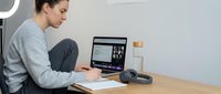 Young Caucasian woman in gray sweater working from home office, taking notes while attending video conference call on laptop computer at wooden desk.