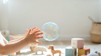 Child hands reaching for floating soap bubble in bright playroom with colorful wooden toys creating magical moment of wonder and discovery.