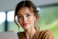Young Caucasian woman with natural makeup and casual brown shirt smiling warmly while working on laptop, soft bokeh background creates professional business atmosphere.