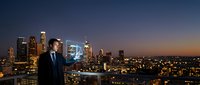 Professional businessman in suit standing on rooftop overlooking illuminated city skyline at dusk with glowing skyscrapers and urban lights below.