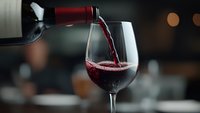 Red wine pouring from bottle into crystal glass with dynamic splash and stream motion, dark atmospheric restaurant background with soft bokeh lights.
