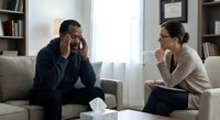 Stressed African American man holding head in hands while Caucasian female therapist provides counseling support in modern office setting.