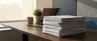 Stack of white paper documents on modern office desk with laptop, coffee mug and plant in background. Professional workspace organization and productivity concept.
