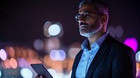 Mature businessman with gray beard and glasses using smartphone at night, illuminated by colorful bokeh city lights in urban environment.