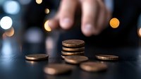 Stacked copper coins on dark surface with blurred bokeh lights and hand reaching in background, representing financial planning and money management.