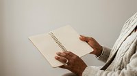 African American person holding open spiral notebook with lined pages, wearing light sweater, planning or writing notes for business, education, or personal journaling purposes.