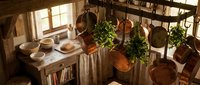 Rustic farmhouse kitchen interior with hanging copper pots, herbs, wooden table and warm sunlight streaming through windows creating cozy cooking atmosphere.