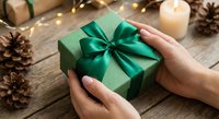 Hands holding green gift box with emerald ribbon on rustic wooden table with candles and pine cones creating festive holiday atmosphere for celebrations.