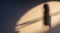 Arched window with dramatic shadows cast on cream wall during golden hour sunlight creating geometric patterns and warm architectural lighting atmosphere.