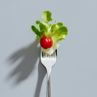 Fresh lettuce leaf and cherry tomato on silver fork against light gray background with dramatic shadow, minimalist food composition for healthy eating concept.