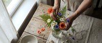 Woman arranging wildflower bouquet with poppies and ferns on rustic wooden table by window. Floral design and home decoration concept.