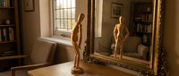 Wooden artist mannequin figure standing on desk beside ornate golden mirror in sunlit study room with books and vintage decor for creative workspace design.