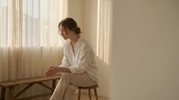 Young woman in white pajamas sitting on wooden stool by window with natural light, peaceful morning routine in minimalist bedroom interior.