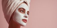 Young woman with facial clay mask and towel wrapped around head against pink background for skincare and beauty concept.