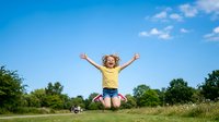 Young girl with blonde hair, approximately 6-8 years old, wearing yellow shirt and denim shorts, jumping with arms raised in celebration outdoors. Green grass field with soccer ball nearby, blue sky with clouds. Caucasian child expressing joy and freedom in nature.