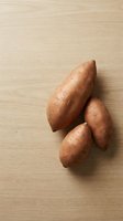 Three fresh sweet potatoes with reddish-brown skin arranged on a light wooden surface, suitable for food photography, cooking blogs, and nutritional content.