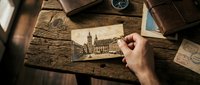 Male hand holding vintage postcard with historic cathedral architecture on rustic wooden desk surrounded by antique travel items, compass, maps and old books for nostalgic exploration themes.