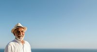 Senior man with light skin tone, wearing white shirt and straw hat with sunglasses, smiling outdoors against blue ocean and sky backdrop for travel, lifestyle, retirement, and wellness concepts.