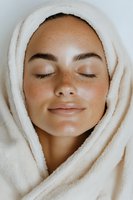Young woman with freckles and closed eyes wrapped in soft white towel, expressing serenity and relaxation during spa treatment or skincare routine.