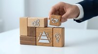 Businessman hand arranging wooden blocks with business icons including gears, handshake, pyramid and lightbulb representing strategy and teamwork concepts.