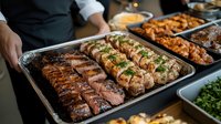 Grilled meat platter with sliced beef steak and chicken breast garnished with fresh herbs on metal catering tray, professional food service setup.