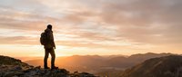 Hiker with backpack standing on mountain peak at sunset, overlooking vast valley landscape during golden hour adventure and outdoor exploration.