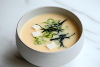 Creamy Asian-style soup with tofu cubes, seaweed strips and green onion garnish in white ceramic bowl on marble surface, soft natural lighting creates appetizing mood.