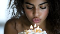 Young mixed-race woman with curly hair blowing out birthday candles on cake, celebrating special occasion with warm candlelight and peaceful expression.