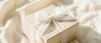 Elegant cream gift box with delicate lace ribbon bow on soft white fabric background for wedding celebrations and special occasions.