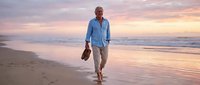 Middle-aged man with gray hair, Caucasian ethnicity, wearing casual blue shirt and beige pants, walking barefoot along sandy beach at sunset holding shoes, peaceful coastal scene.