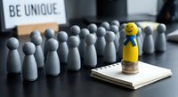 Yellow wooden peg figure with blue ribbon stands out from crowd of gray pegs on desk with be unique sign and notepad for leadership concepts.