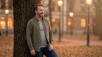 Mature Caucasian man in casual attire leaning against tree in autumn park at dusk, warm street lights glowing in background, contemplative mood for lifestyle and seasonal themes.