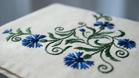 Delicate blue floral embroidery with green vine scrollwork on cream linen fabric showcasing traditional needlework craftsmanship for textile and handicraft projects.