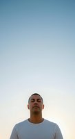 Clear sky portrait of a man with short dark hair and olive complexion wearing a light gray t-shirt, looking upward against a gradient blue and white sky at dusk or dawn.