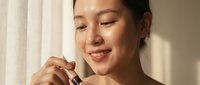 Young Asian woman applying facial serum with dropper in natural sunlight by window, skincare routine morning beauty ritual self-care wellness.