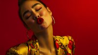 Young Asian woman with red lipstick and gold hoop earrings in yellow floral dress poses sensually against red background, eyes closed in dramatic lighting.
