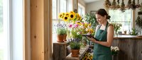 Young female florist arranging fresh sunflowers and lavender in rustic flower shop interior with natural lighting and wooden decor elements.