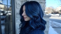 Young Asian woman with vibrant blue wavy hair styled in loose curls wearing knit sweater, side profile view showcasing modern hair coloring and styling technique.