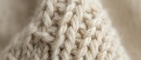 Close-up macro shot of cream colored knitted wool fabric showing detailed cable stitch pattern texture for cozy winter fashion and craft projects.