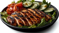 Grilled chicken breast with char marks served on fresh green salad with cherry tomatoes and cucumber slices on black plate, close up view of protein meal.