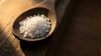 Coarse sea salt crystals in rustic wooden spoon on dark wood surface for cooking, seasoning and culinary applications in kitchen and restaurant use.