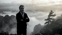 Confident businessman standing on mountain peak above clouds with arms crossed, dramatic sky and rocky landscape background for success concepts.